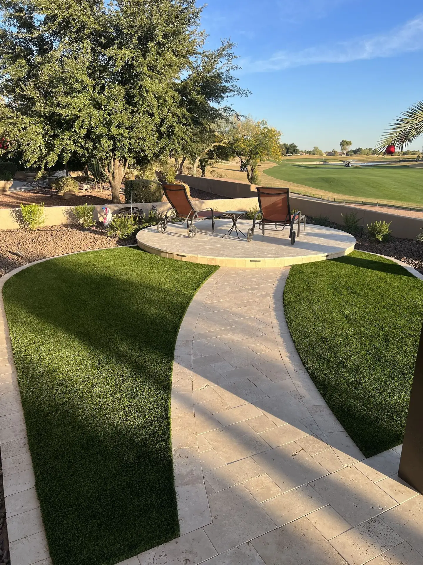 Artificial turf and travertine paver walkway in modern front yard design, featuring lounge chairs and lush greenery, enhancing curb appeal and inviting atmosphere.