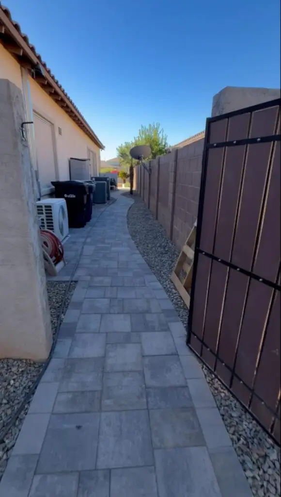 Paved walkway leading to a gated entrance, flanked by a wall and greenery, showcasing a well-maintained outdoor space in a residential area.