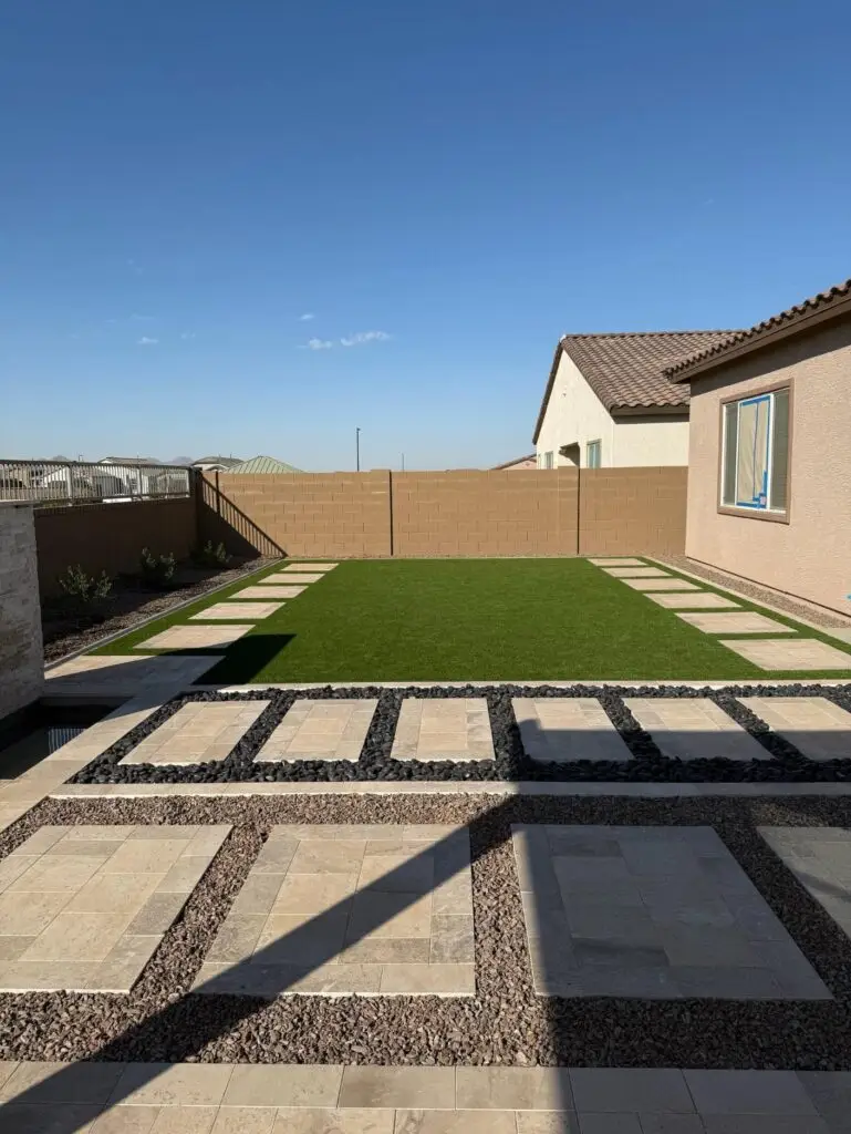 Backyard remodel in Queen Creek, AZ featuring travertine pavers, artificial turf, and decorative gravel pathways under a clear blue sky, creating a functional and inviting outdoor space.
