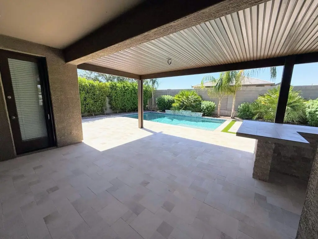 Backyard remodel in Queen Creek, AZ, featuring travertine pavers, outdoor kitchen under pergola, lush greenery, and a swimming pool, creating a vibrant and functional outdoor space.