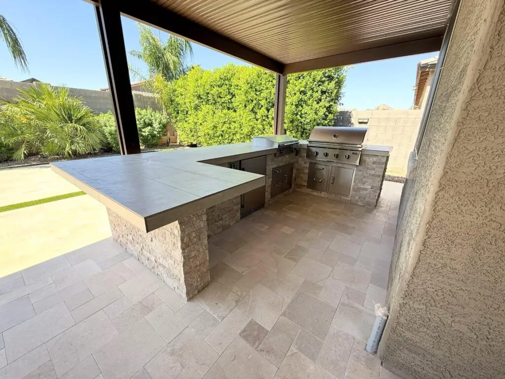 Outdoor kitchen with modern grill and countertop, surrounded by lush greenery and travertine pavers, enhancing backyard functionality and aesthetics in Queen Creek, AZ.