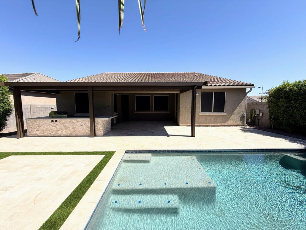 Backyard remodel in Queen Creek, AZ, featuring a modern outdoor kitchen, travertine pavers, artificial turf, and a swimming pool, designed for relaxation and entertainment under a pergola.