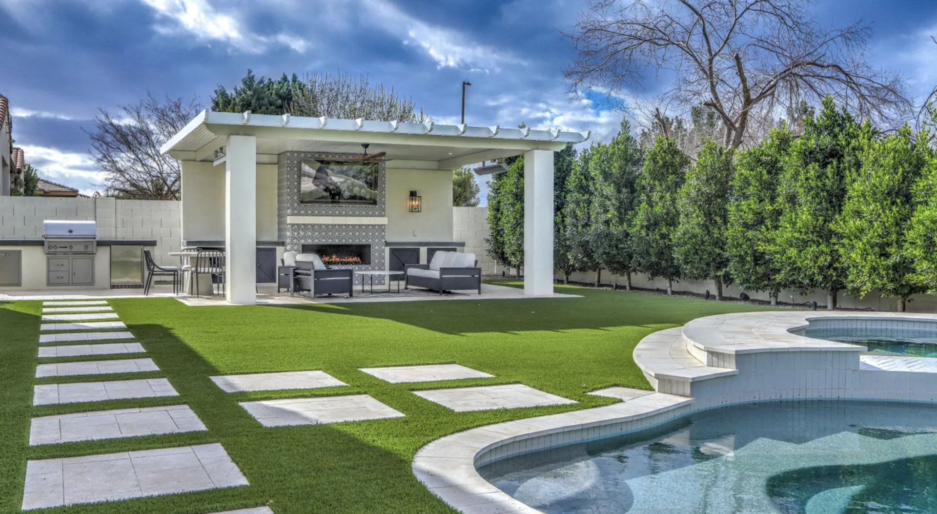 Full backyard remodel in Chandler, AZ with a remodeled pool, white ramada with fireplace, artificial turf lawn and travertine stepping pads