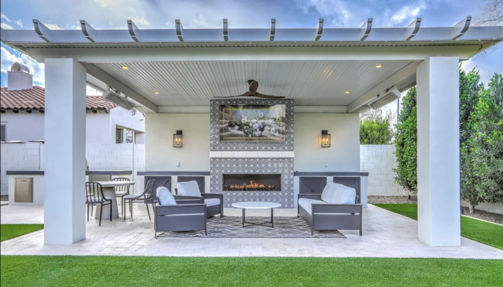Covered ramada in a Chandler, AZ backyard with a linear gas fireplace, tile surround, mounted television and travertine paver patio